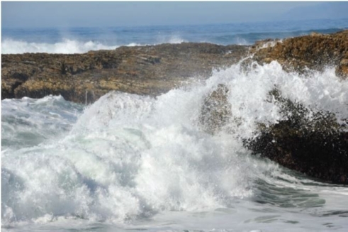 Below is a picture of water striking rocks at a coastline. Describe which of Earth's spheres are interacting here, and some of the effects that may be caused by this intera