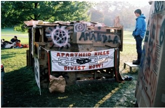 <strong>The image Antiapartheid Protest, Cornell University from 1986 shows students with a self-made shantytown hut resembling those in South Africa. What does this image suggest?  </strong> A) American businesses employed South African workers under unspeakable conditions. B) American businesses were involved in South African businesses. C) Cornell students had brought their own experiences in South Africa home. D) Cornell University was using South Africans for unethical research and experiments. <div style=padding-top: 35px> 